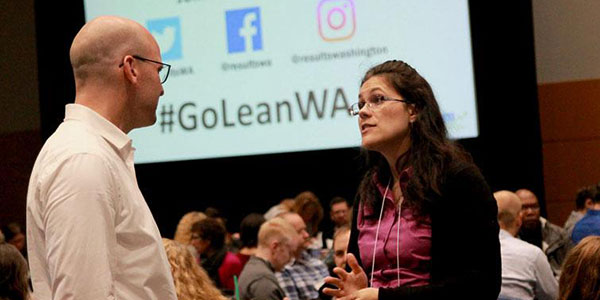 Two attendees at the Washington State Government Lean Transformation conference sharing ideas between break out sessions. In the background and center of photo there's a projection screen with the hashtag #GoLeanWa along with Twitter, Facebook, and Instagram Icons with a message that says "Join the conversation."