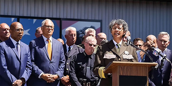 Governor Inslee stands with a group of officers and government leaders behind a police officer standing at a podium who appears to be making a speech. 