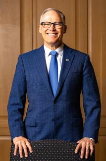 Governor Jay Inslee, a male with grey hair and glasses in a blue suit, stands smiling behind a chair in a wood-paneled room
